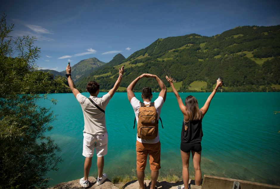Three young adults celebrating by a vibrant turquoise lake surrounded by lush hills.