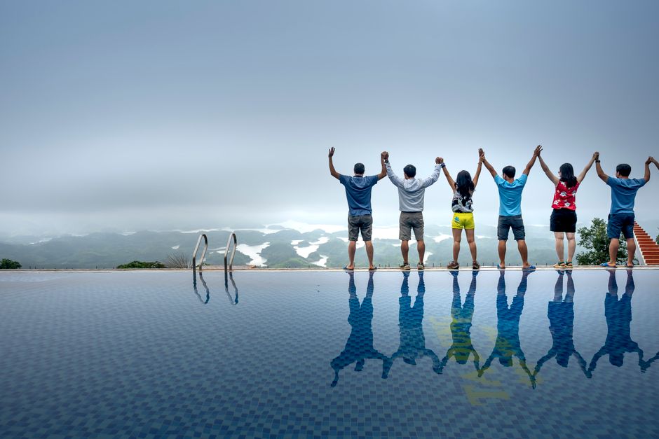 Seven people standing by an infinity pool with arms raised, reflected in water, overlooking scenic foggy landscape.