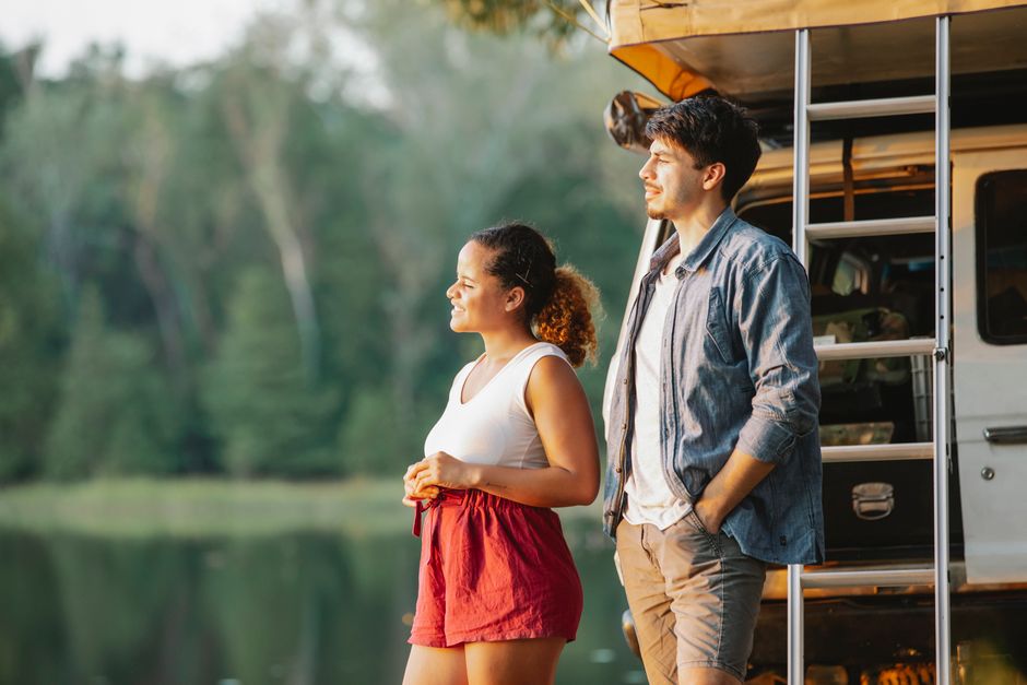 Multiracial couple near SUV car on lake shore during road trip at sunset.